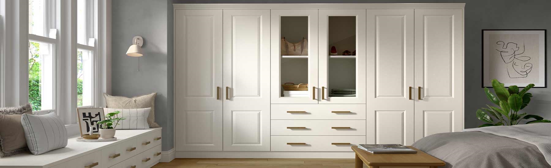 Spacious bedroom with white wardrobes, wooden handles, and glass panels. Light wood floor, fluffy rug, and a cozy vibe with a small side table and soft lighting.