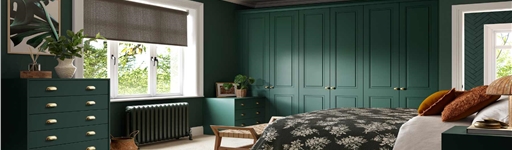 Cozy bedroom with dark green walls and furniture, a floral bedspread, woven rug, and a large window allowing natural light. Calm and inviting atmosphere.