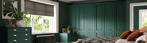 Cozy bedroom with dark green walls and furniture, a floral bedspread, woven rug, and a large window allowing natural light. Calm and inviting atmosphere.