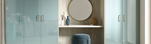 A modern bedroom features glossy pastel blue wardrobes with integrated vanity. A round mirror, decorative vases, and a soft velvet stool complete the elegant look.