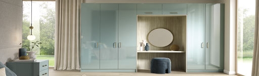 A modern bedroom features glossy pastel blue wardrobes with integrated vanity. A round mirror, decorative vases, and a soft velvet stool complete the elegant look.