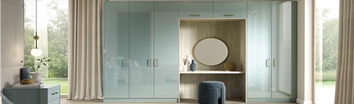 A modern bedroom features glossy pastel blue wardrobes with integrated vanity. A round mirror, decorative vases, and a soft velvet stool complete the elegant look.