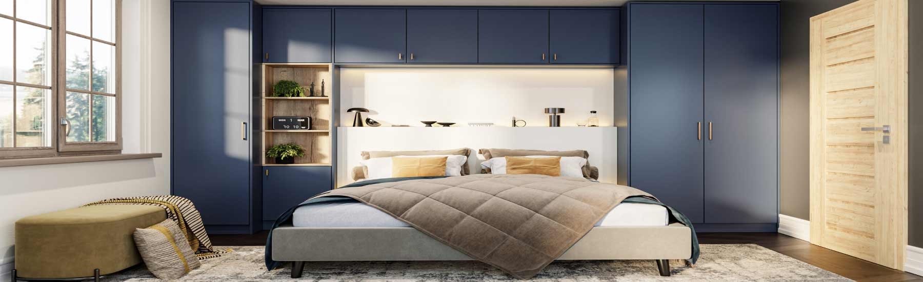 An image of a classic cream-coloured fitted bedroom wardrobe installation surrounding a double bed. The built-in unit features floor-to-ceiling storage, including tall double wardrobes on either side of the bed and a row of four overhead cupboards bridging across the top. The doors have a traditional Shaker style with subtle vertical grooved panelling and are fitted with small, round wooden knobs. The matching cream furniture extends to a bedside chest of drawers, a blanket box at the foot of th