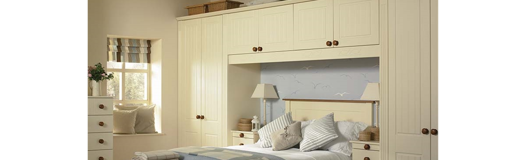 An image of a classic cream-coloured fitted bedroom wardrobe installation surrounding a double bed. The built-in unit features floor-to-ceiling storage, including tall double wardrobes on either side of the bed and a row of four overhead cupboards bridging across the top. The doors have a traditional Shaker style with subtle vertical grooved panelling and are fitted with small, round wooden knobs. The matching cream furniture extends to a bedside chest of drawers, a blanket box at the foot of th