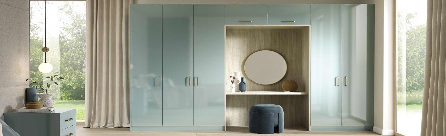 A modern bedroom features glossy pastel blue wardrobes with integrated vanity. A round mirror, decorative vases, and a soft velvet stool complete the elegant look.